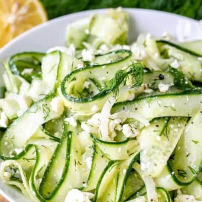 Cucumber Feta Salad in a white bowl, with dill and lemon in the background