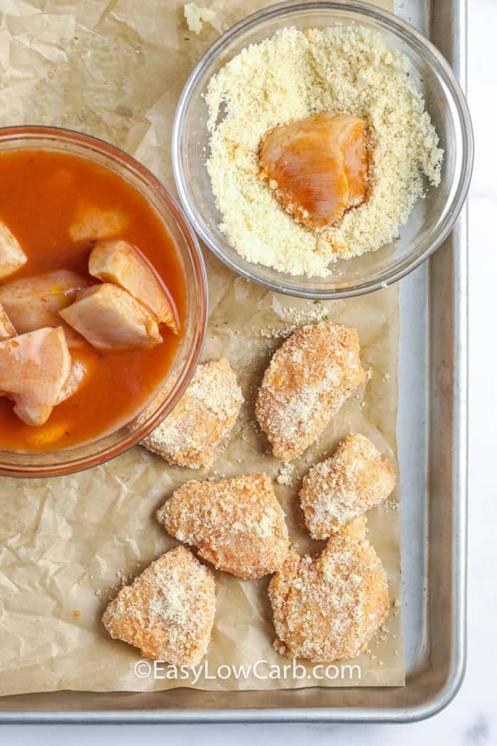 Chicken marinated in a bowl of buffalo sauce, then dipped in a bowl of coating and placed on a lined baking sheet.