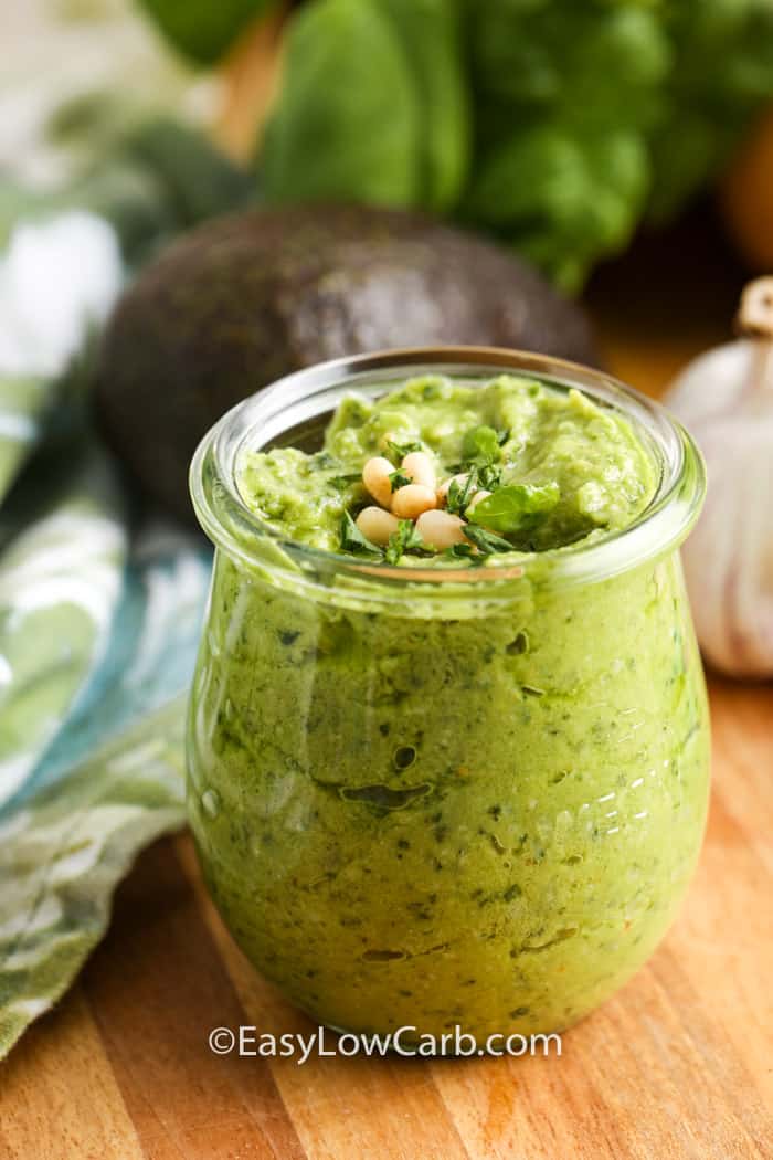 This keto avocado pesto sauce is a bright and creamy way to add flavor to low carb meals. Fresh basil, avocado, garlic, lemon juice, parmesan, pine nuts, and olive oil create a smooth sauce that works with warm or chilled dishes. Try it with zoodles, grilled chicken, salmon, roasted vegetables, or as a flavorful spread for low carb wraps. #easylowcarb Avocado Pesto in a jar with ingredients around it