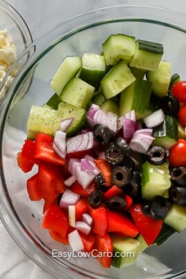 greek salad ingredients in a clear bowl with feta cheese on the side in a small clear bowl
