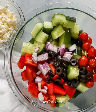 greek salad ingredients in a clear bowl with feta cheese on the side in a small clear bowl