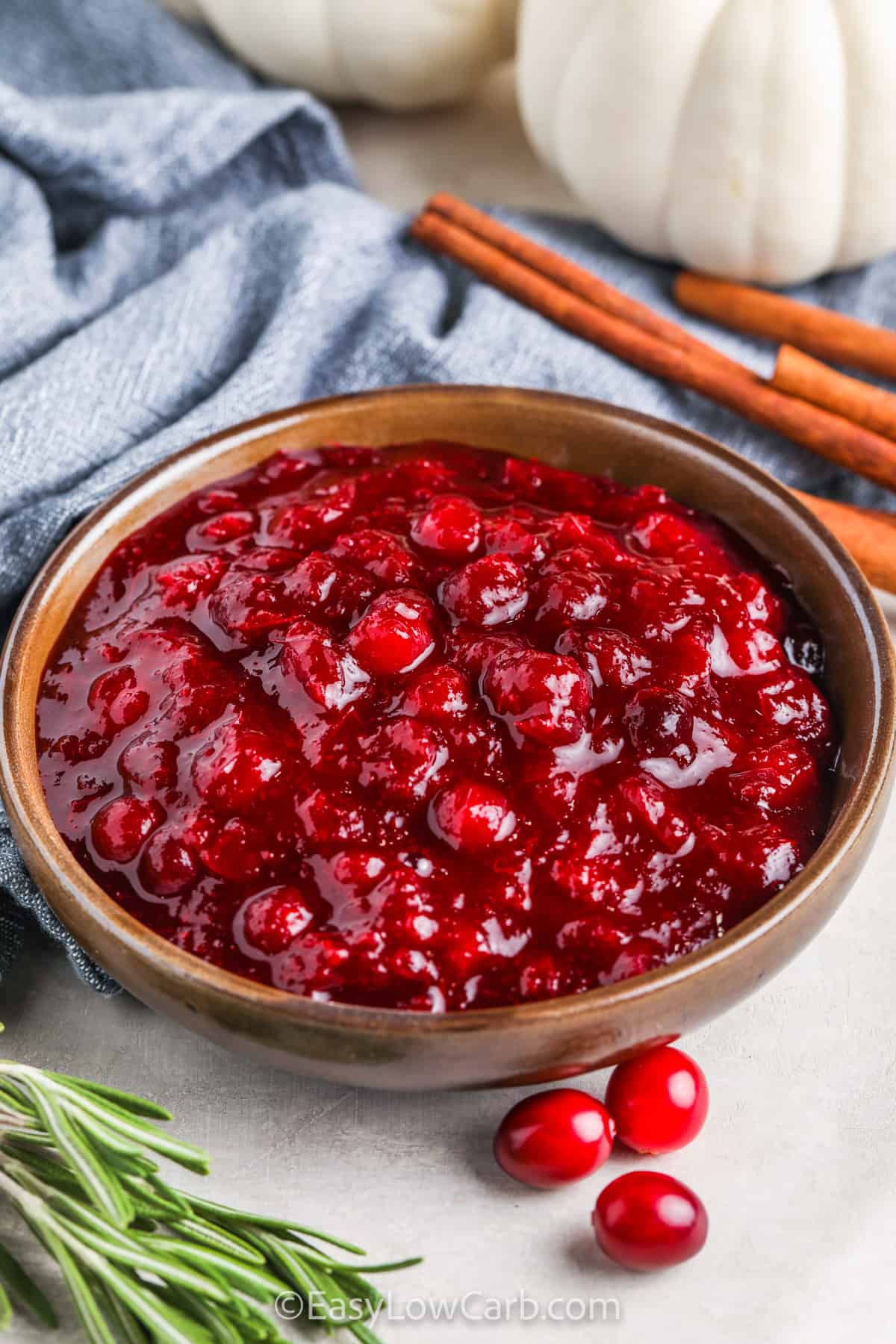 Keto Cranberry Sauce in a bowl with cranberries, rosemary and cinnamon sticks on the side