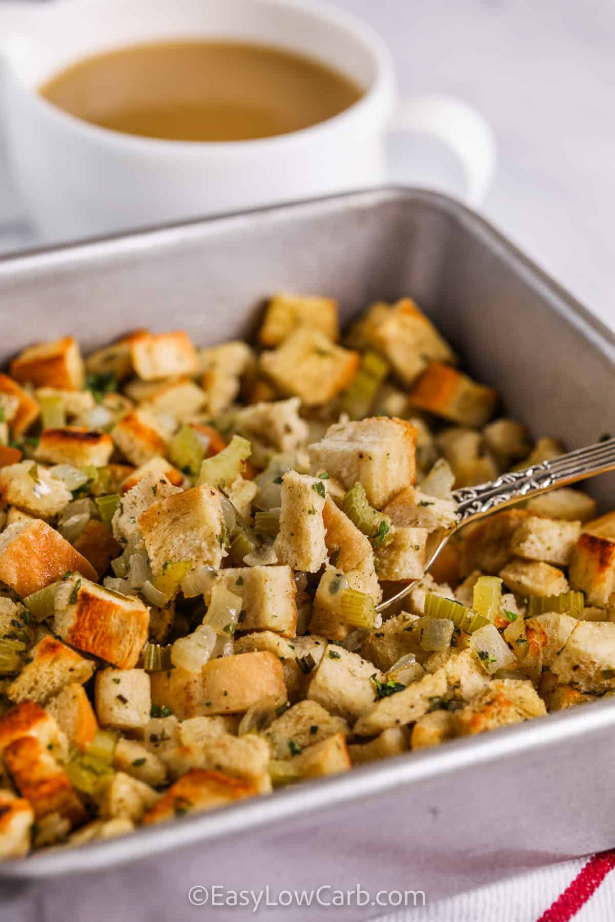 Keto stuffing combines crisp toasted bread cubes, sautéed onions, celery, poultry seasoning, and fresh herbs. Savory chicken broth keeps it moist while baking to a golden finish. This low carb side is simple, easy, and perfect to make for Thanksgiving or any holiday gathering. It’s a flavorful, aromatic dish that captures the warmth and nostalgia of classic stuffing while staying light and healthier for everyone to enjoy. #easylowcarb taking a spoonful of Keto Stuffing from the pan