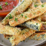 close up of baked keto zucchini fries piled on plate with a small bowl of marinara sauce in the background