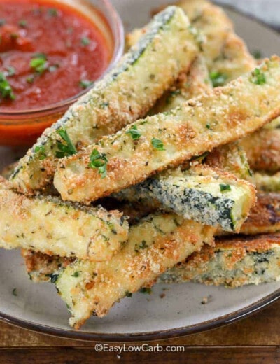 close up of baked keto zucchini fries piled on plate with a small bowl of marinara sauce in the background