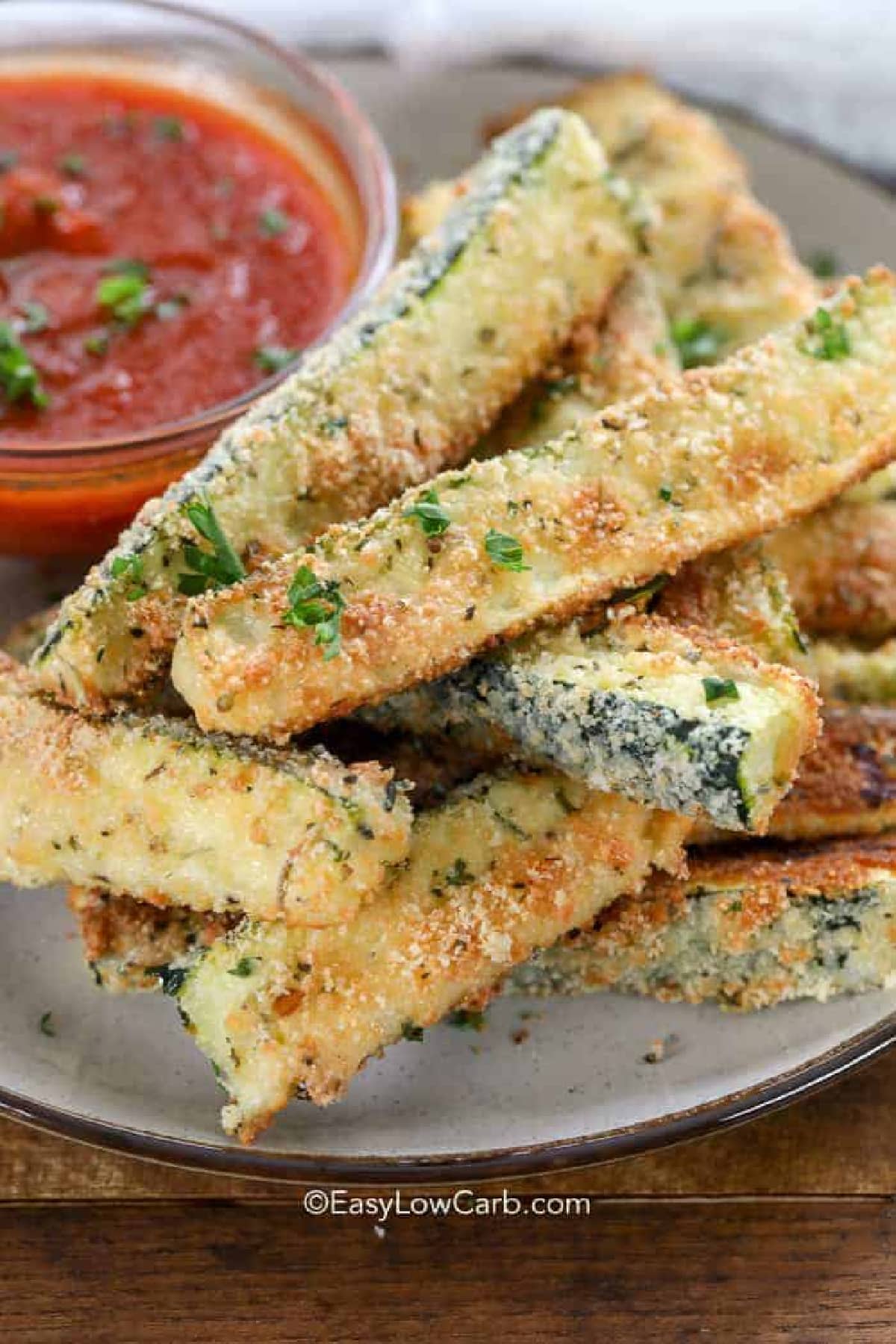 close up of baked keto zucchini fries piled on plate with a small bowl of marinara sauce in the background