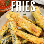 baked keto zucchini fries piled on plate with a small bowl of marinara sauce in the background, with a title