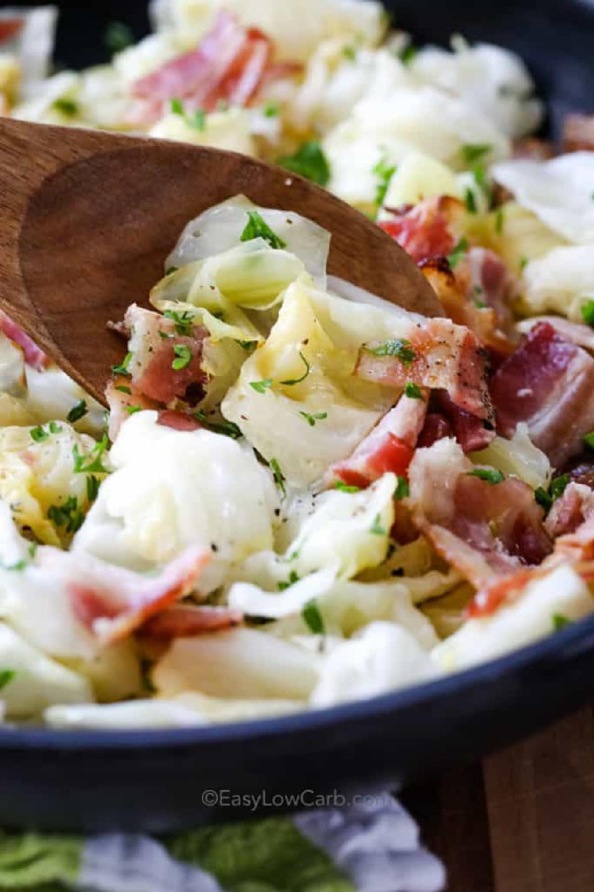 Fried cabbage with bacon in a skillet and stirred with a wooden spoon