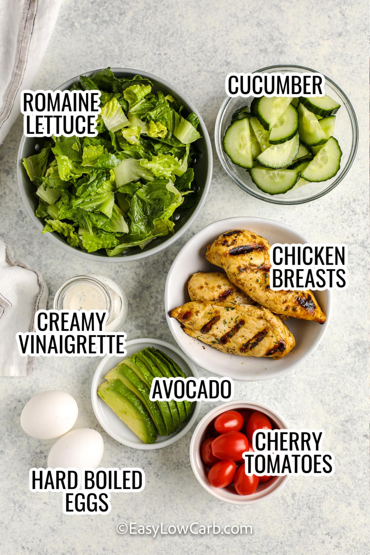 ingredients assembled to make grilled chicken salad, including romaine lettuce, cucumber, creamy vinaigrette, avocado, cherry tomatoes, chicken breasts, and hard boiled eggs