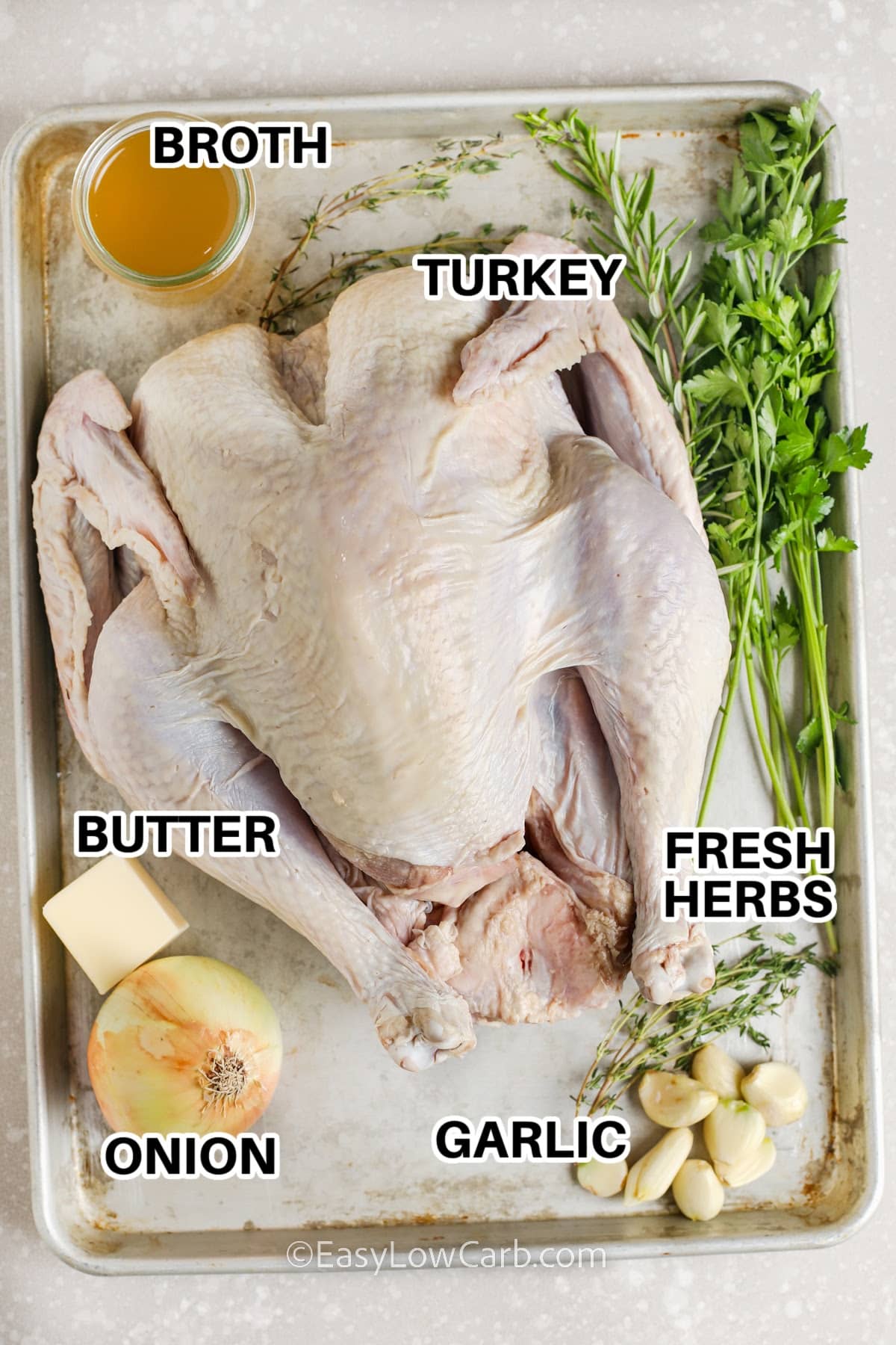 ingredients to make oven roasted turkey on a baking tray labeled: turkey, broth, butter, onion, garlic, and fresh herbs