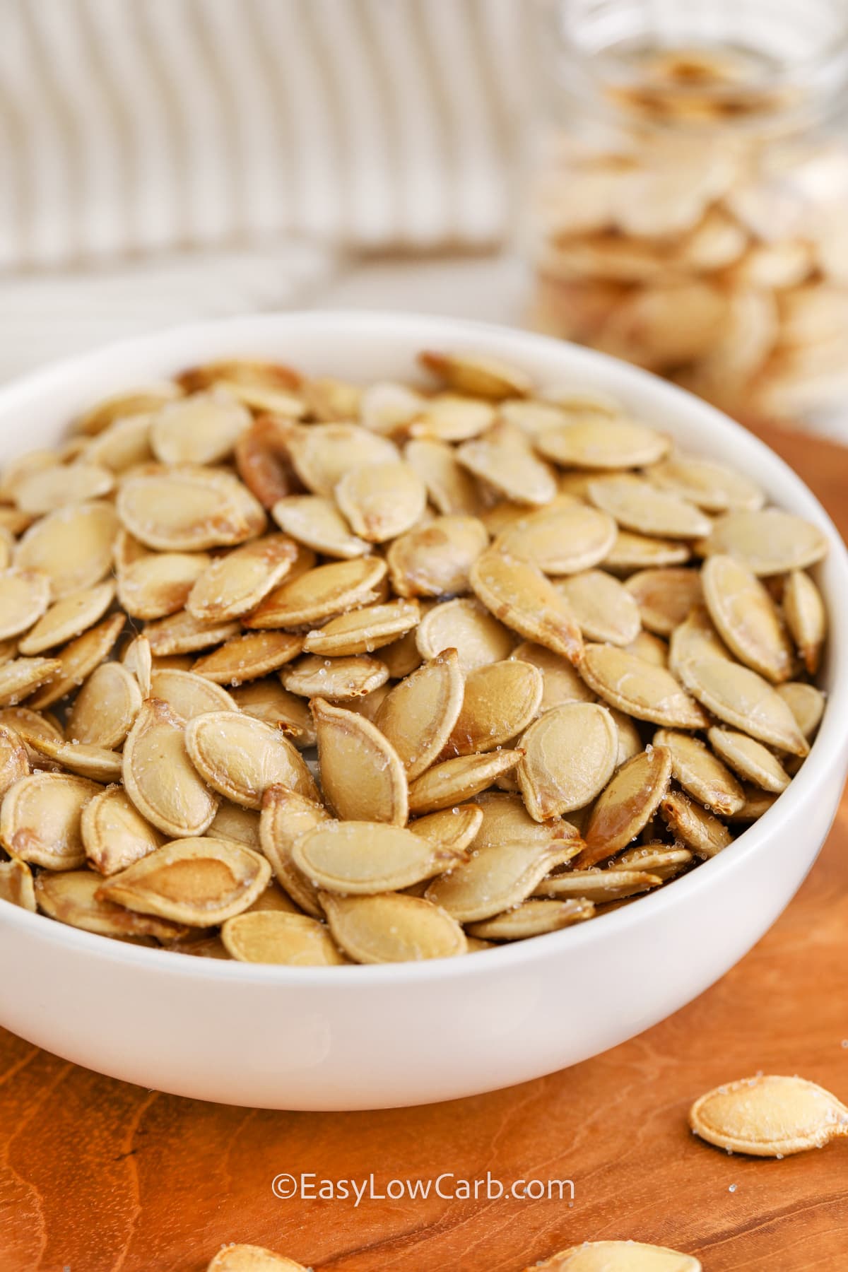 Baked pumpkin seeds are an easy, homemade snack that’s perfect for the Thanksgiving or Halloween season. These crunchy seeds are tossed with olive oil and salt for a savory and salty flavor that complements any autumn gathering. Their roasted, nutty taste makes them a wholesome treat you can enjoy anytime. Customize them with spices for a sweet or spicy twist to match your seasonal cravings. #easylowcarb close up of baked pumpkin seeds in a white bowl