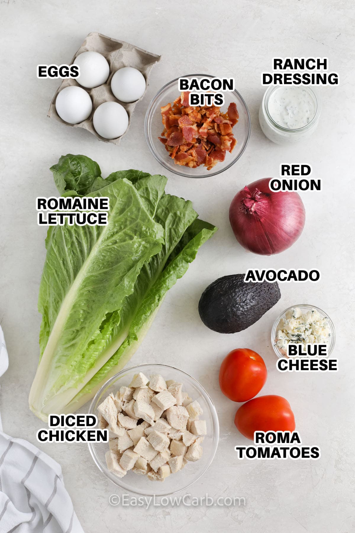 Cobb Salad Recipe ingredients including eggs, bacon bits, ranch dressing, red onion, avocado, blue cheese, roma tomatoes, diced chicken, and romaine lettuce