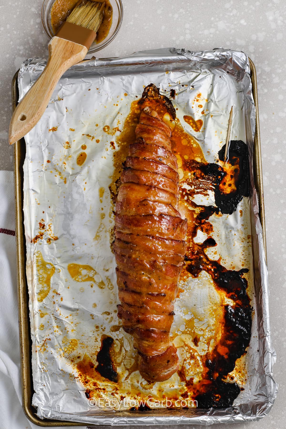 bacon wrapped pork tenderloin cooked on a baking sheet with glaze brushed on top.