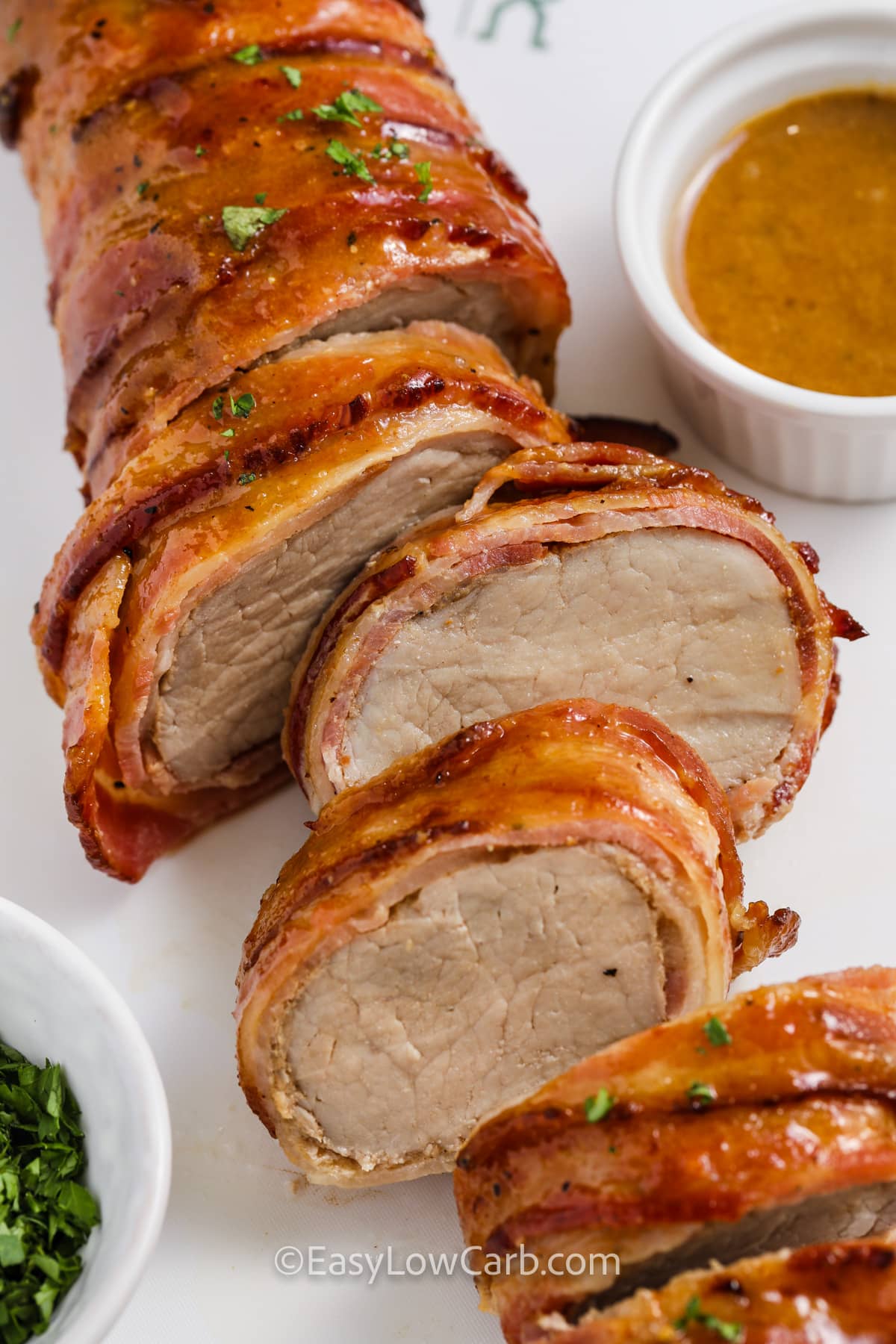 This bacon wrapped pork tenderloin recipe uses a simple glaze made with Dijon mustard, brown sugar sweetener, salt, and pepper. The bacon wraps the pork to protect it from drying out, and the glaze adds a caramelized, savory-sweet flavor. Once baked to temperature, brush on the pan-juice glaze and let it rest, then slice for an easy low carb main dish. #easylowcarb sliced bacon wrapped pork tenderloin
