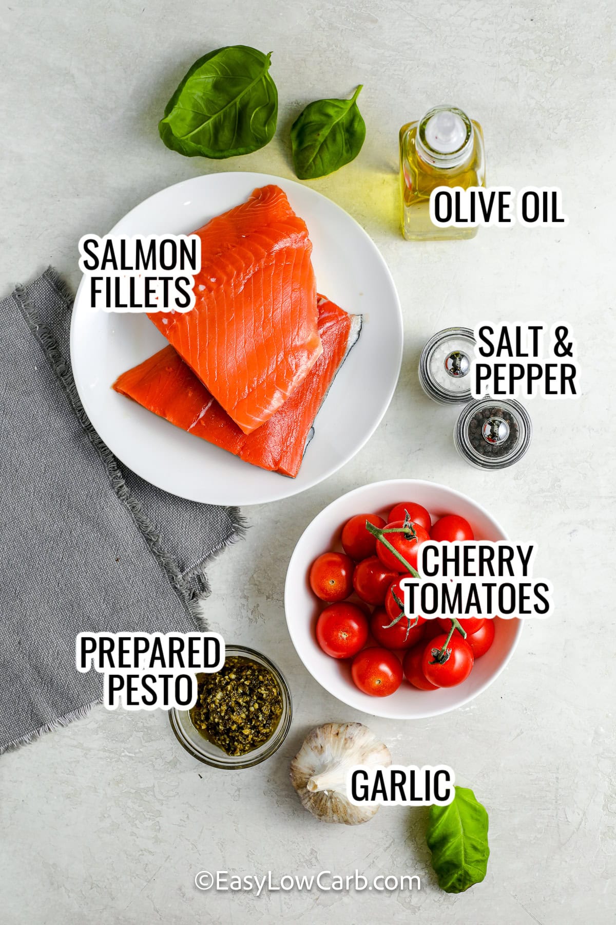 ingredients assembled to make pesto salmon, including salmon fillets, olive oil, cherry tomatoes, pesto, and garlic