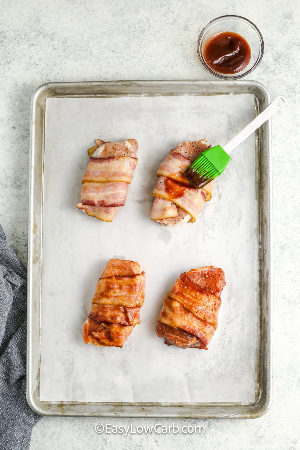 Four bacon wrapped pork chops being brushed with barbecue sauce on a baking tray