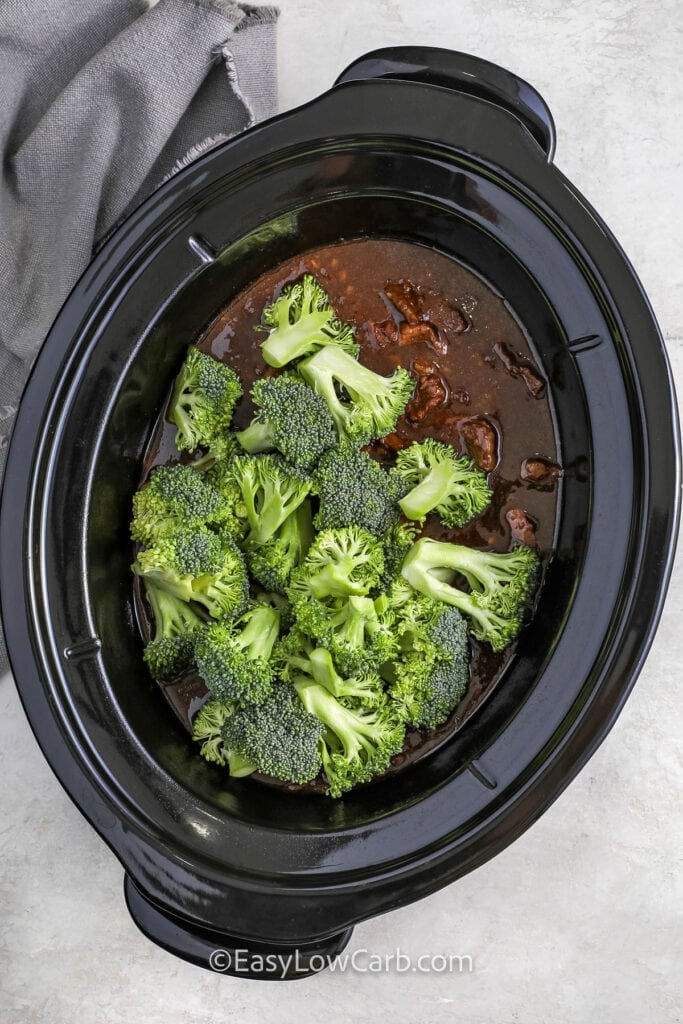 Adding broccoli florets to a crock pot of slow cooker beef and broccoli.