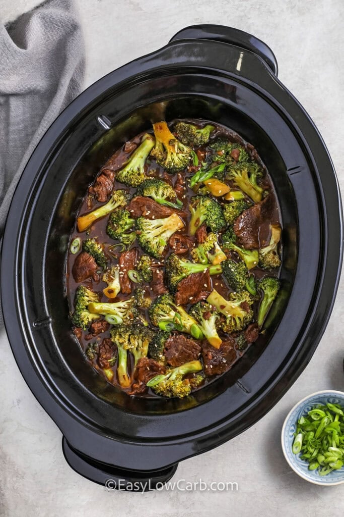 Looking down on a crock pot of cooked slow cooker beef and broccoli with a bowl of green onions.