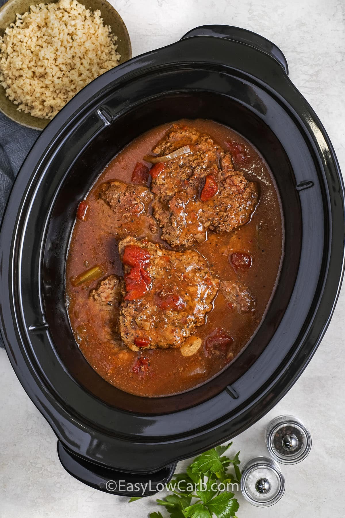 slow cooker cube steak in a crock pot with cauliflower rice, salt and pepper in the background