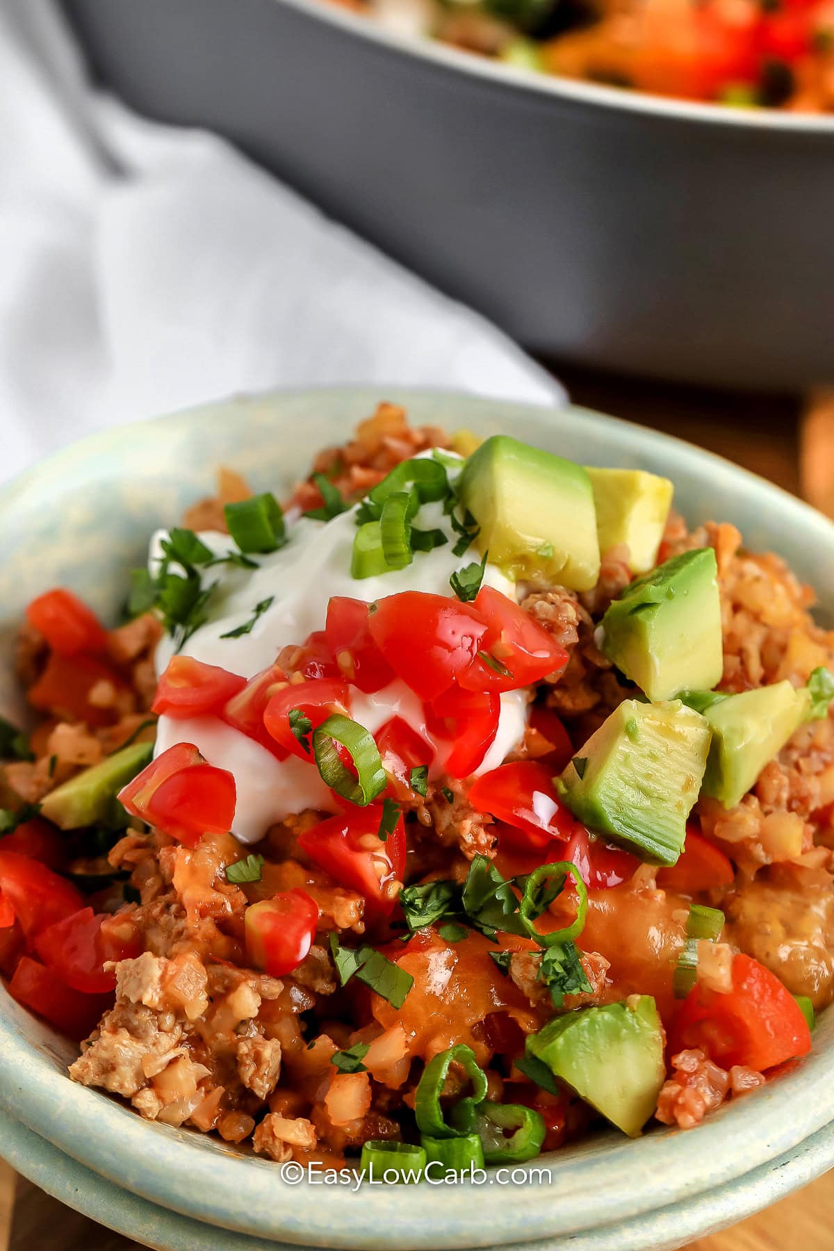This cheesy taco skillet is one-pan dinner packed with bold flavors. Ground beef, cauliflower rice, and colorful bell peppers are simmered with taco seasoning and salsa for a savory base. Top it off with melty cheese and fresh toppings like avocado, sour cream, and tomatoes for a customizable meal. Ready in under 30 minutes, it’s an easy weeknight meal. #easylowcarb bowl of Cheesy Taco Skillet with pan in the back