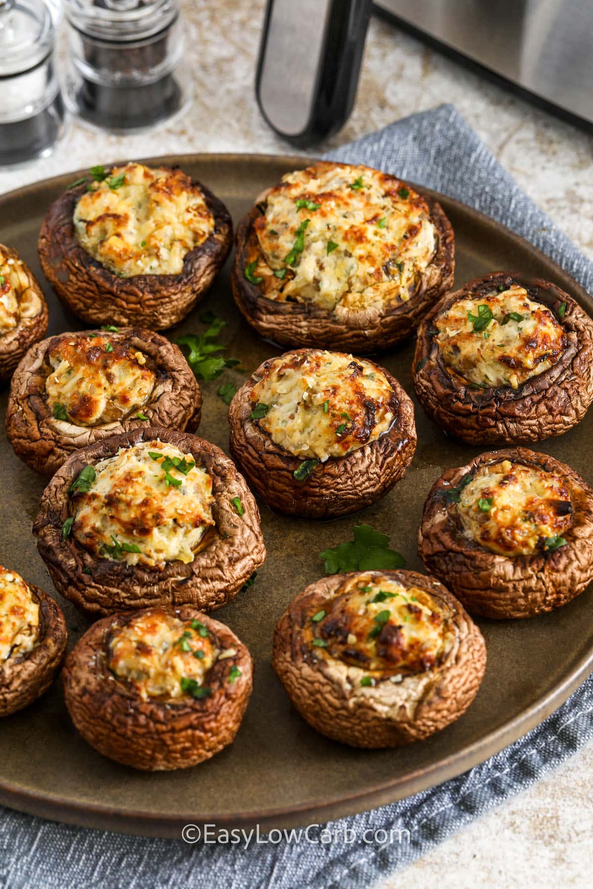 Air fryer stuffed mushrooms are the ultimate bite-sized snack that’s packed with bold flavor and cheesy goodness. The mushrooms are stuffed with a creamy blend of cream cheese, garlic, parmesan, and fresh herbs, then cooked in the air fryer until golden and bubbly. The result is a crispy, savory, and absolutely delicious appetizer that’s perfect for any occasion. They take just minutes to prepare and make an excellent low-carb alternative to traditional party snacks. #easylowcarb close up of Air Fryer Stuffed Mushrooms on a plate