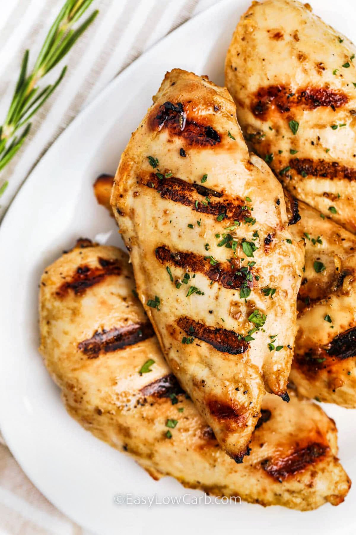 Grilled chicken breasts are made for summer cooking, especially when marinated in a zesty blend of garlic, lemon, mustard, and spices. They grill up juicy and golden every time, making them one of the easiest and most flavorful recipes to keep on hand. Perfect for a quick weeknight dinner or low carb meal prep, this high-protein dish is always a hit. Pair it with grilled veggies or a fresh salad. #easylowcarb close up of Grilled Chicken Breasts on a plate
