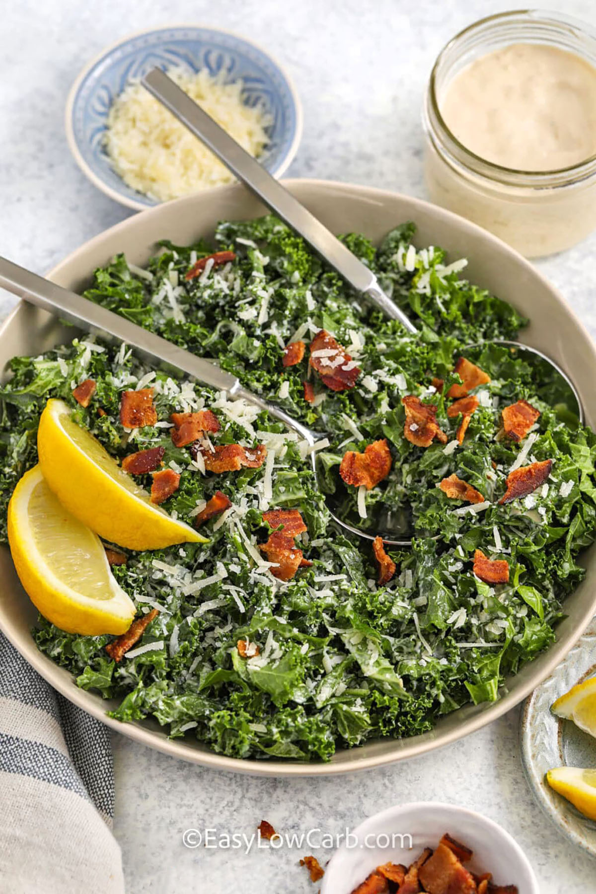 Kale Caesar salad is a flavorful, low-carb salad that keeps its crisp texture in the dressing. Massaged kale, crisp bacon, and Parmesan cheese combine for a savory bite with a bright touch from lemon wedges. Top with chicken or salmon to make a complete main dish. This salad brings fresh flavor and satisfying crunch to lunches, dinners, or meal prep. It is an easy way to enjoy a healthy dish without sacrificing taste or richness in every bite. #easylowcarb bowl of Kale Caesar Salad with spoons