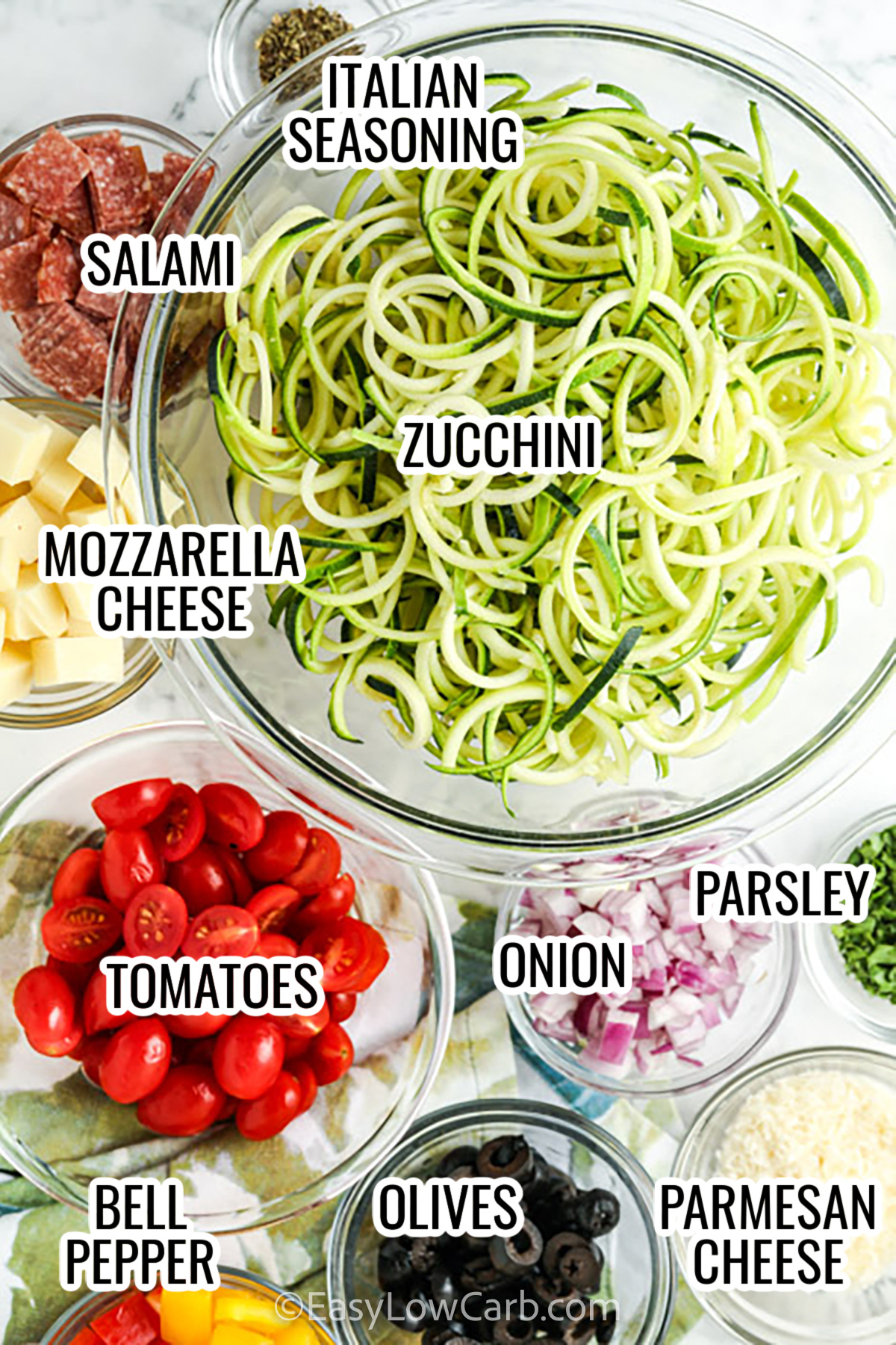 ingredients assembled to make zucchini pasta salad, including zucchini, olives, salami, mozzarella cheese, tomatoes, parmesan cheese, bell peppers, onion, and italian seasoning