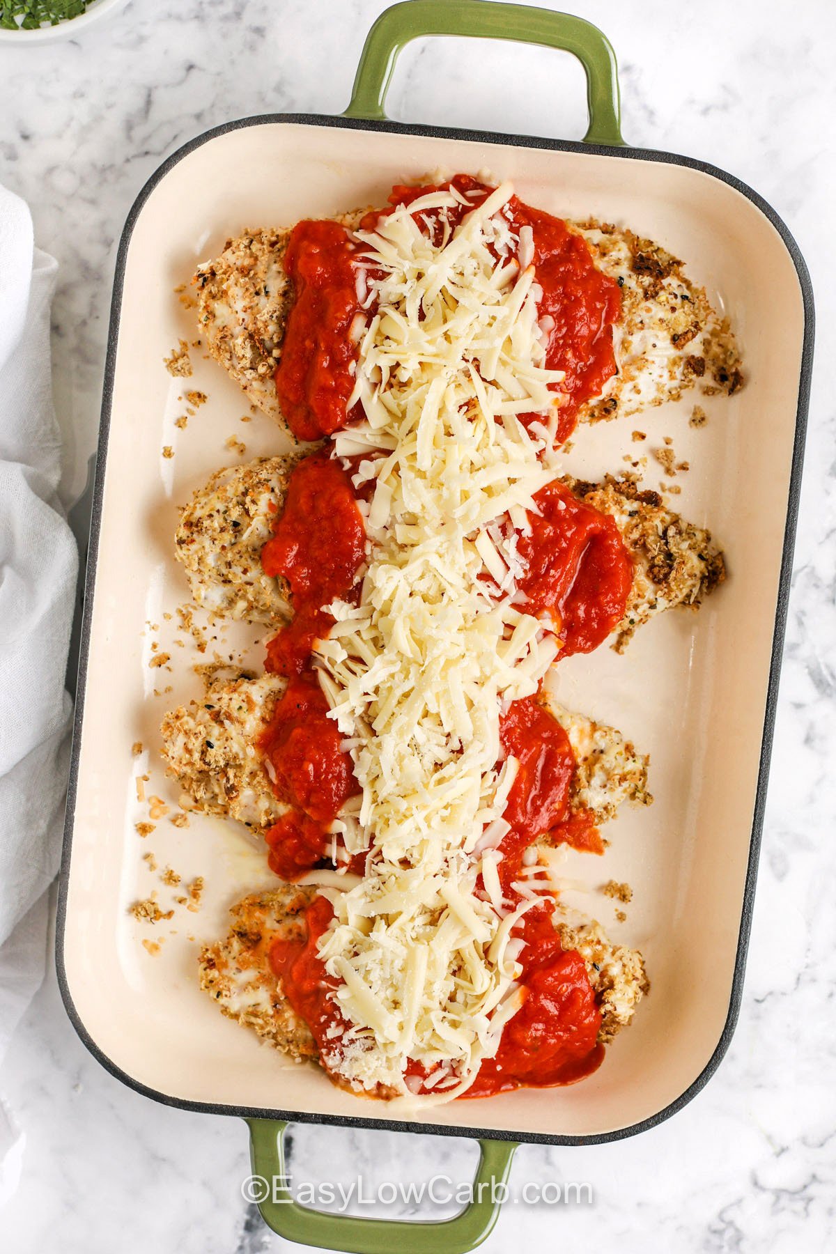 This oven baked chicken Parmesan starts with tender chicken cutlets coated in a mix of Parmesan cheese, Italian seasoning, and keto breadcrumbs for that perfect golden crunch. After baking, it’s topped with rich, flavorful pasta sauce and a blanket of melted mozzarella. The result is a warm, cheesy, satisfying dish that’s lighter, healthier, and every bit as delicious as the traditional fried version. #easylowcarb adding sauce and cheese on top of breaded chicken to make Oven Baked Chicken Parmesan