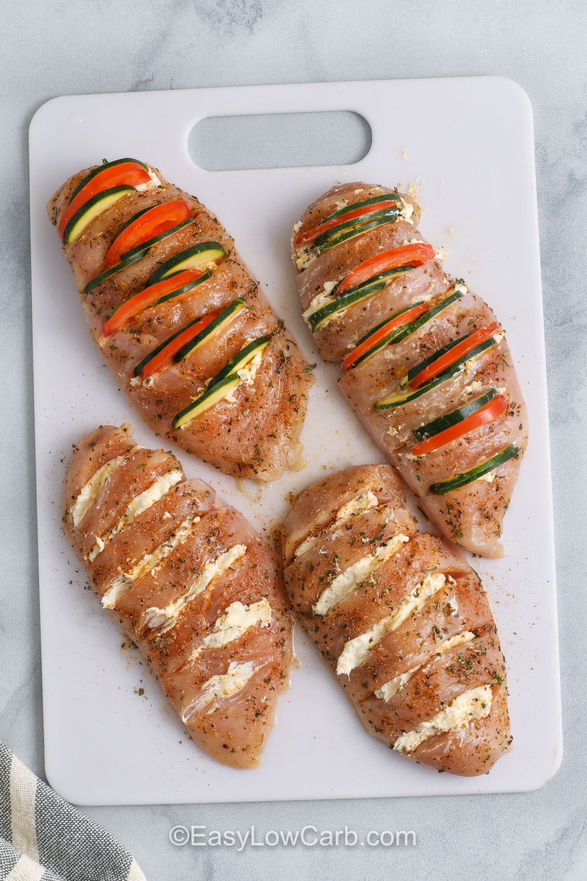 Hasselback chicken transforms plain chicken breasts into a cheesy, veggie-stuffed delight. Each cut is filled with a blend of cream cheese, lemon zest, and tender slices of zucchini and tomato. It’s low in carbs, high in flavor, and perfect for impressing guests without much effort. This baked dish comes together fast, and the oven does all the hard work for you. Easy, healthy, and delicious! #easylowcarb hasselback chicken being prepared on a cutting board