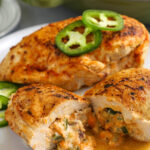 two jalapeno popper chicken breasts on a plate topped with sliced jalapenos with one cut in half and the filling melting out