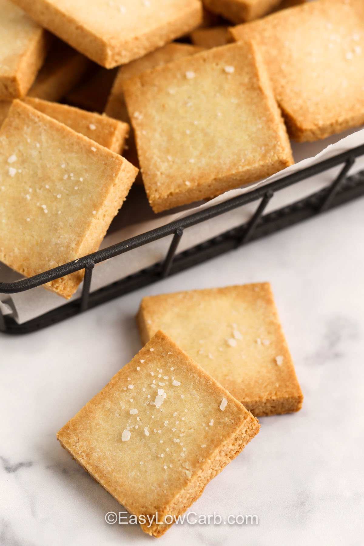 This keto crackers recipe is your new go-to for crunchy, low-carb snacks! Made with almond flour and seasoned simply with garlic powder and kosher salt, they’re easy to prepare and totally satisfying. No need to buy store-bought when homemade tastes this good. Roll out, cut, bake, and enjoy in no time. Great for lunchboxes or charcuterie boards! #easylowcarb two prepared keto crackers on the counter with a basket of keto crackers behind them
