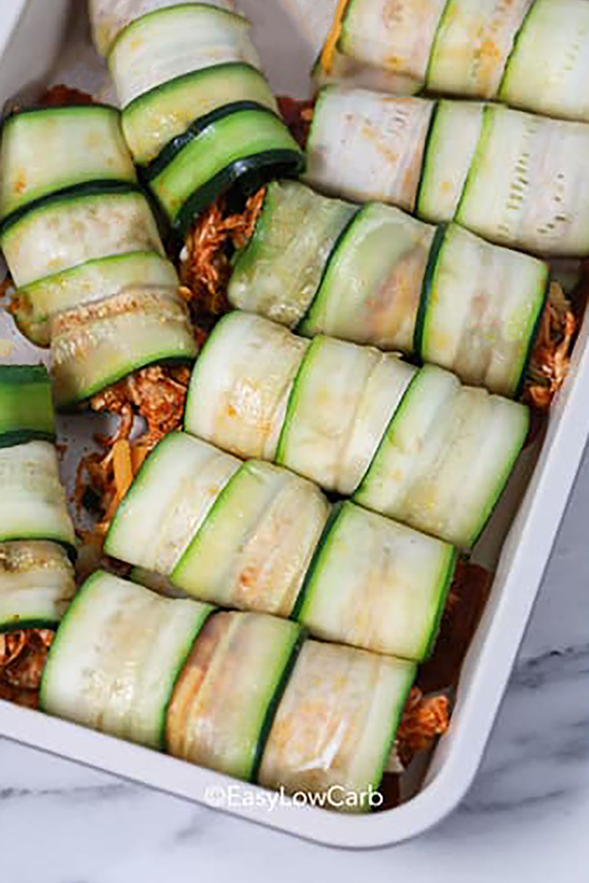 For light eating with big flavor, try low carb chicken zucchini enchiladas. It’s the perfect recipe for extra zucchini and leftover chicken. Use a vegetable peeler or mandoline to shave zucchini into flexible ribbons that hold a hearty chicken and cheese filling. Enchilada sauce keeps everything moist and adds bold flavor, while the topping of cheddar and Monterey Jack browns nicely in the oven. #easylowcarb adding Zucchini Enchiladas to the dish before baking