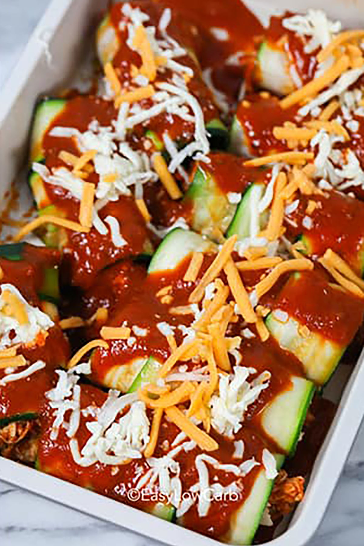 For light eating with big flavor, try low carb chicken zucchini enchiladas. It’s the perfect recipe for extra zucchini and leftover chicken. Use a vegetable peeler or mandoline to shave zucchini into flexible ribbons that hold a hearty chicken and cheese filling. Enchilada sauce keeps everything moist and adds bold flavor, while the topping of cheddar and Monterey Jack browns nicely in the oven. #easylowcarb adding sauce and cheese to Zucchini Enchiladas before cooking