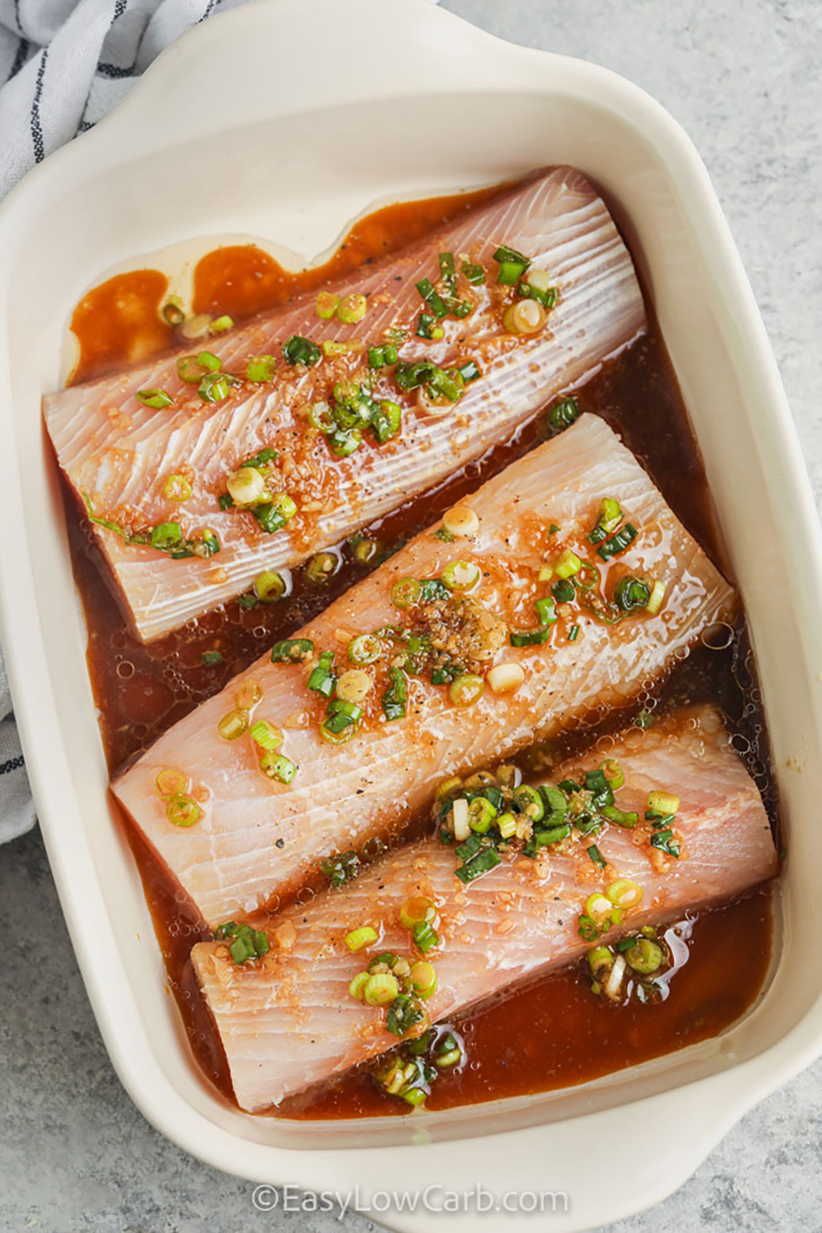 This oven baked mahi mahi is mild, meaty, and perfect for a bold sesame ginger marinade. Soy sauce and lime create a savory, bright glaze that coats the fish as it bakes. Use an instant-read thermometer to avoid overcooking and keep the texture moist. Top with green onions and sesame, then serve with cauliflower rice to soak up the sauce. #easylowcarb mahi mahi in a casserole dish with sauce , ginger , garlic and green onions to make Baked Mahi Mahi Recipe