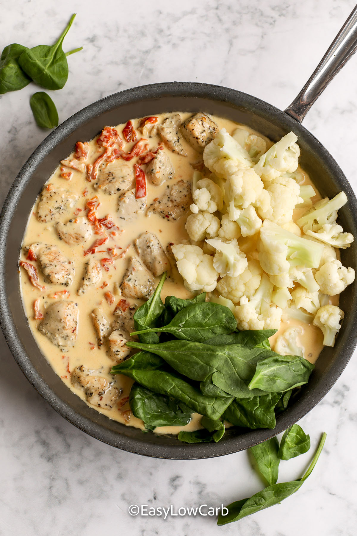 Low carb creamy Tuscan chicken skillet is a cozy comfort meal with chicken, spinach, and sun-dried tomatoes in creamy garlic Parmesan sauce. Par-cooking the cauliflower in broth adds flavor and helps prevent watery sauce. Finish everything together in the skillet until hot and glossy. #easylowcarb preparing tuscan chicken in a skillet with basil, cauliflower chicken and sauce