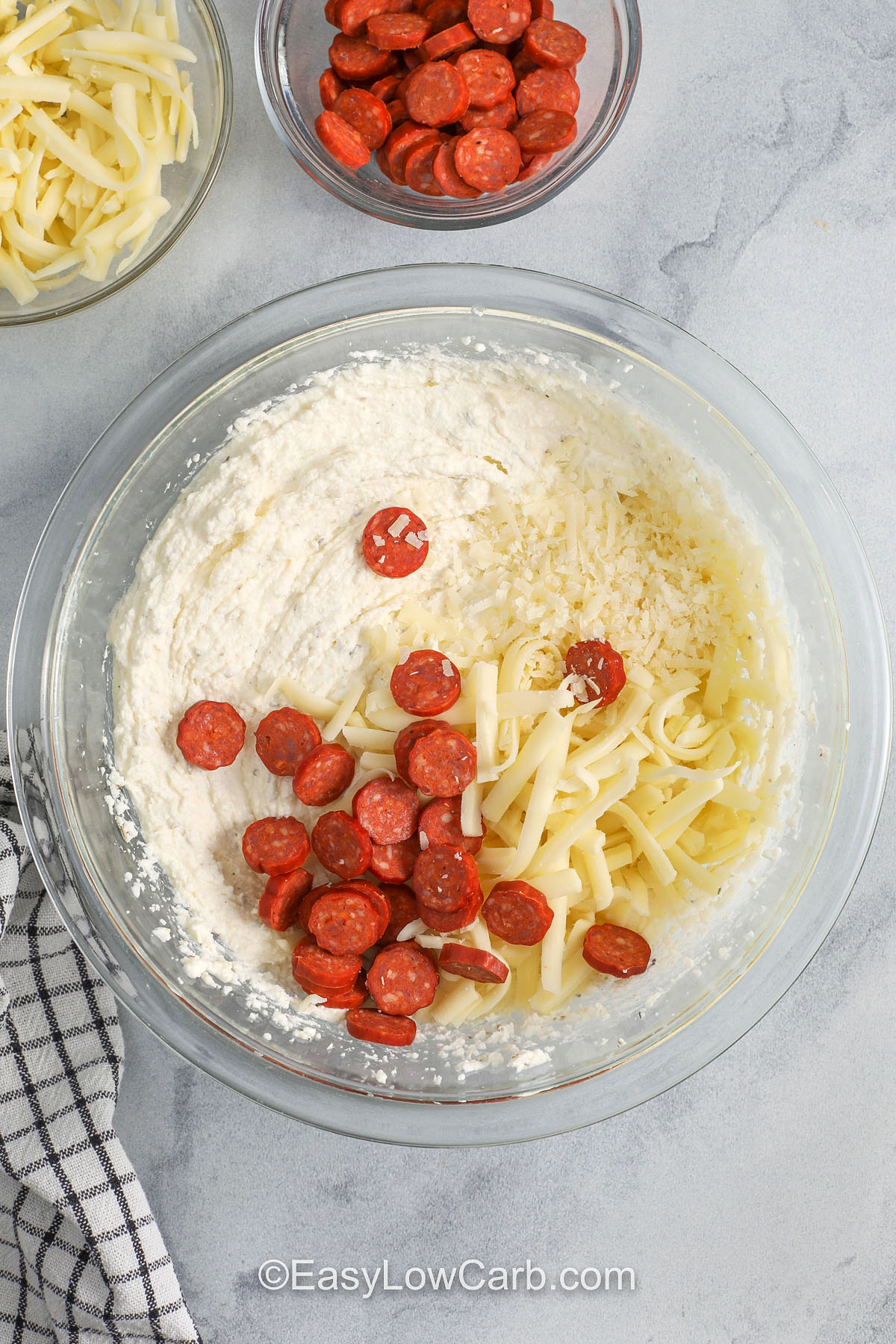 This low carb pepperoni dip is rich, cheesy, and made for sharing. Mix some mozzarella and pepperoni into the whipped ricotta base so every scoop has flavor, then top with marinara, more cheese, and pepperoni. Bake until bubbly and lightly browned. Use a low sugar marinara and shred your own mozzarella for an extra stretchy, melty finish. #easylowcarb adding cheese and pepperoni to bowl to make Pepperoni Dip