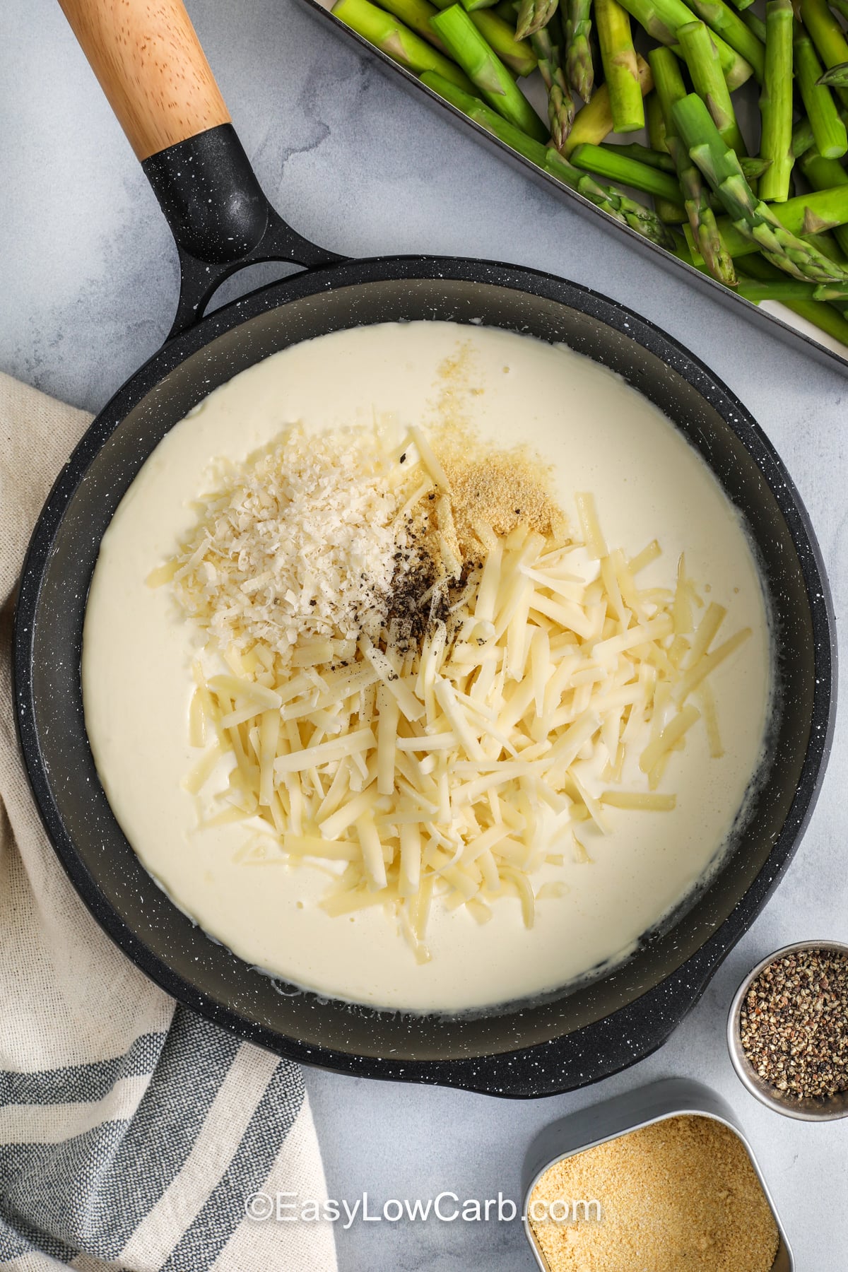 This cheesy asparagus casserole is a simple way to turn fresh asparagus into a crowd-pleasing side. A quick stovetop sauce with garlic, cream cheese, and heavy cream turns smooth and rich, then cheddar and parmesan melt in for deep flavor. Sprinkle crushed pork rinds on top for a crisp, browned finish. It is keto-friendly, gluten free, and great for meal prep. #easylowcarb adding cheese and seasonings to ingredients in pan to make sauce for Asparagus Casserole
