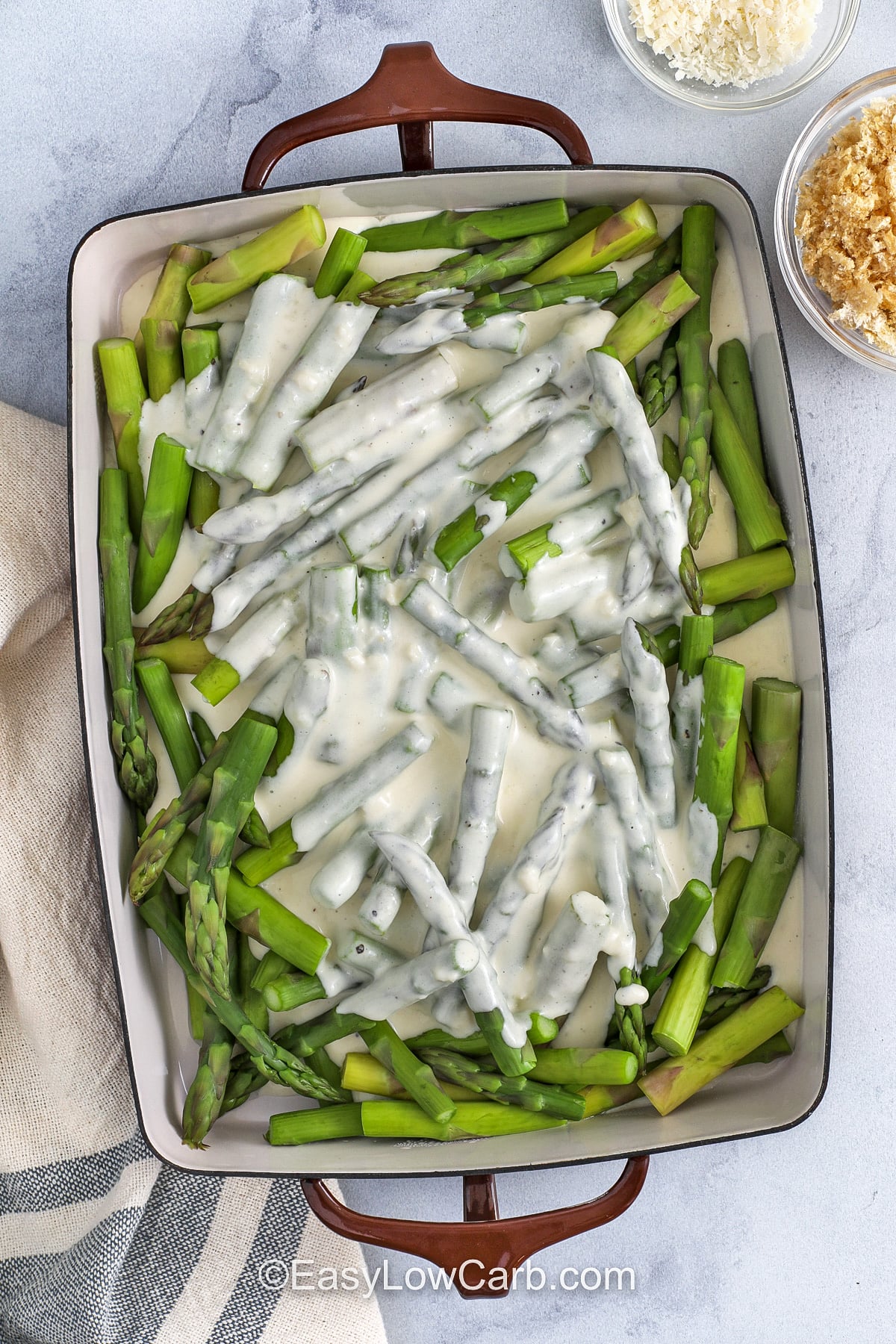 If you love cheesy vegetable bakes, this asparagus casserole delivers a creamy middle and a crispy top in one easy dish. Blanching the asparagus first keeps it from turning soft in the oven. The sauce is made with heavy cream and cream cheese, then finished with cheddar and parmesan for a savory bite. Crushed pork rinds replace breadcrumbs for a low carb crunch. #easylowcarb adding sauce on top of asparagus to make Asparagus Casserole