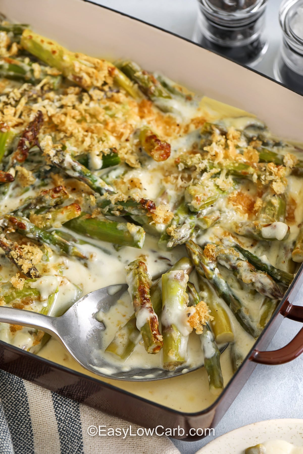 This asparagus casserole is a creamy, cheesy side dish with tender-crisp asparagus baked in a garlic cream cheese sauce. Heavy cream keeps the texture rich while sharp cheddar or gruyere adds bold flavor. A crunchy topping of crushed pork rinds and parmesan gives you that classic casserole finish while keeping it low carb and gluten free. Serve it with steak, pork chops, or chicken for an easy keto-friendly dinner. #easylowcarb creamy Asparagus Casserole in a dish with a spoon