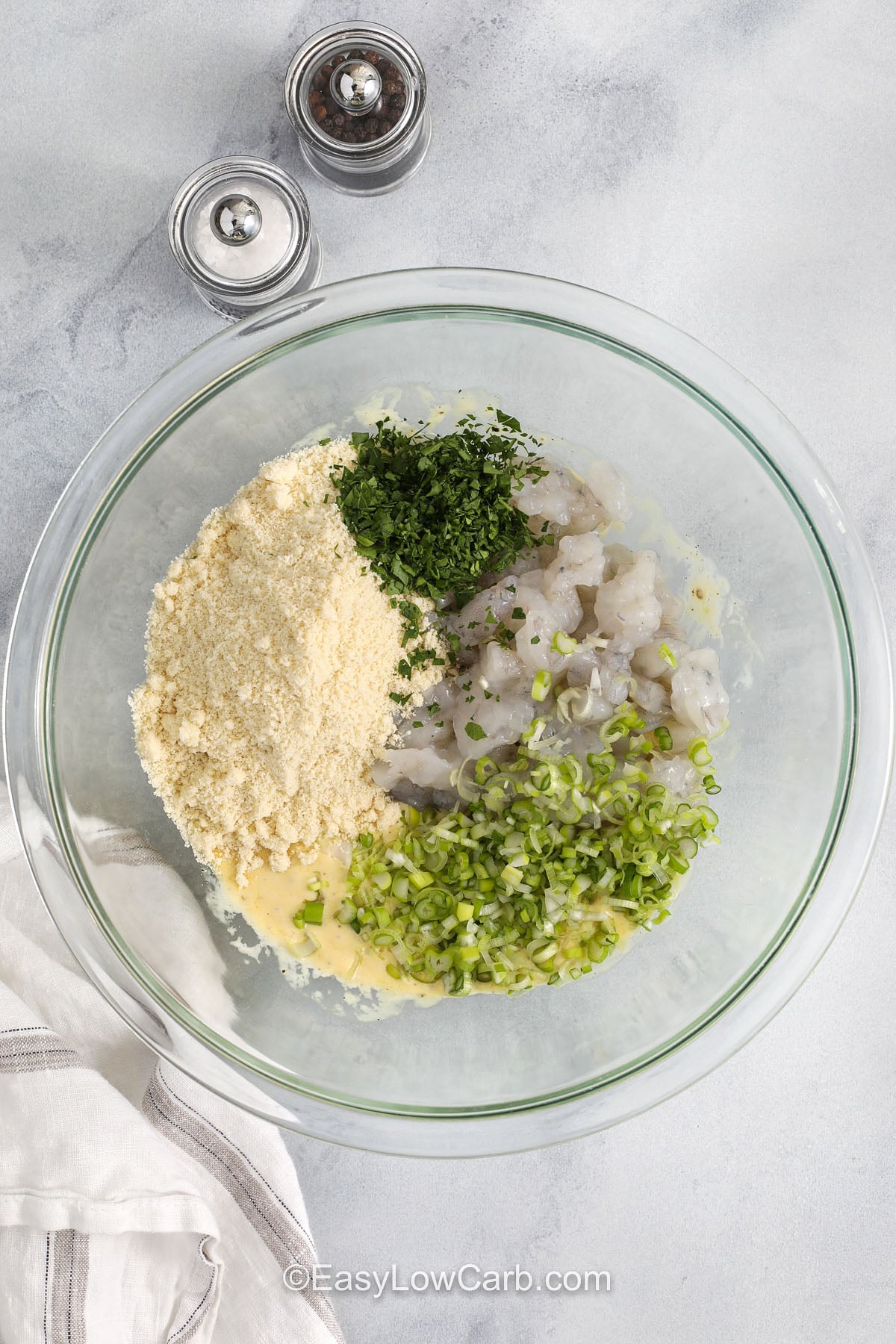 These shrimp cakes are a simple skillet recipe that turns raw shrimp, Dijon, parsley, and parmesan into a crisp, flavorful meal. Finely chopped shrimp gives them a tender texture, while almond flour keeps them low carb and helps them hold together. Serve these shrimp cakes with lemon aioli and a fresh salad for an easy dinner that feels polished but still practical for a busy night. #easylowcarb adding ingredients to bowl to make Shrimp Cakes
