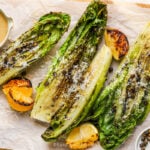 Grilled romaine lettuce and lemon on a a cutting board with a homemade dressing and capers to the side.