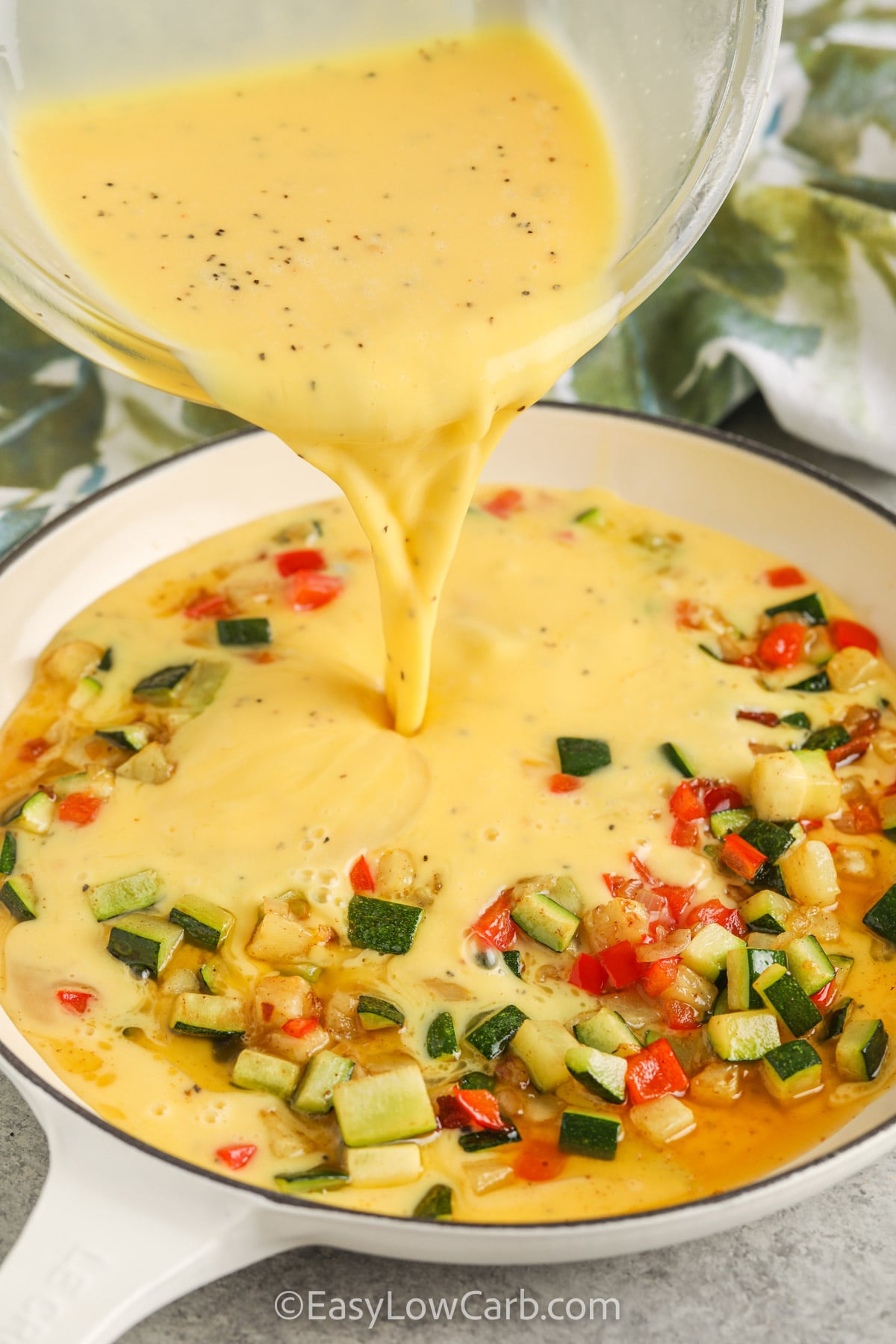 Zucchini frittata is the kind of easy breakfast recipe you'll want to make again and again. Crispy bacon, tender zucchini, sweet bell pepper, and cheddar cheese bake into every flavorful bite. This low carb frittata is great for meal prep, weekend brunch, or breakfast for dinner. It is hearty enough to keep everyone happy and simple enough for busy days. Try this zucchini egg bake today. #easylowcarb pouring egg over veggies to make Zucchini Frittata