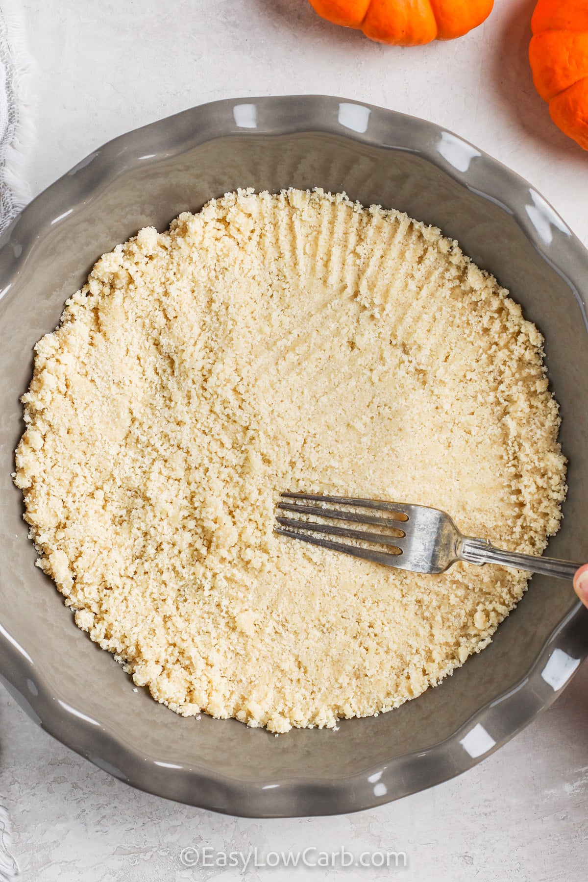 This keto pumpkin cheesecake recipe is a simple way to put a rich fall dessert on the table without a traditional graham cracker crust. Finely ground almond flour, butter, and keto sweetener form the base, while pumpkin puree and cream cheese create a thick, creamy filling. The cheesecake needs several hours to chill, making it an excellent dessert to prepare the night before. Save it for an easy make-ahead Thanksgiving treat. #easylowcarb flattening crust to dish to make a Keto Pumpkin Cheesecake
