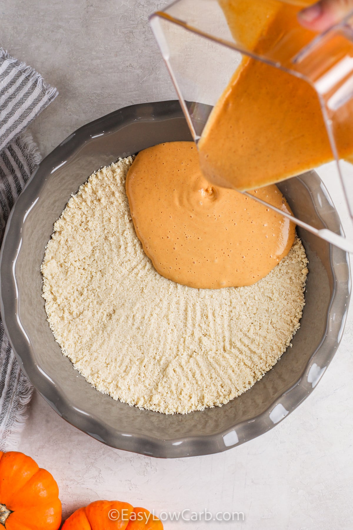 Keto pumpkin cheesecake is rich enough for a holiday table but simple enough to prepare without a springform pan or water bath. The pumpkin and cream cheese filling is blended until smooth, poured over a lightly browned almond flour crust, and baked before chilling until firm. Serve it cold with whipped cream or chopped pecans for extra texture. Save this creamy low carb pumpkin dessert for Thanksgiving, Christmas, or any fall gathering. #easylowcarb pouring Keto Pumpkin Cheesecake mix over crust.