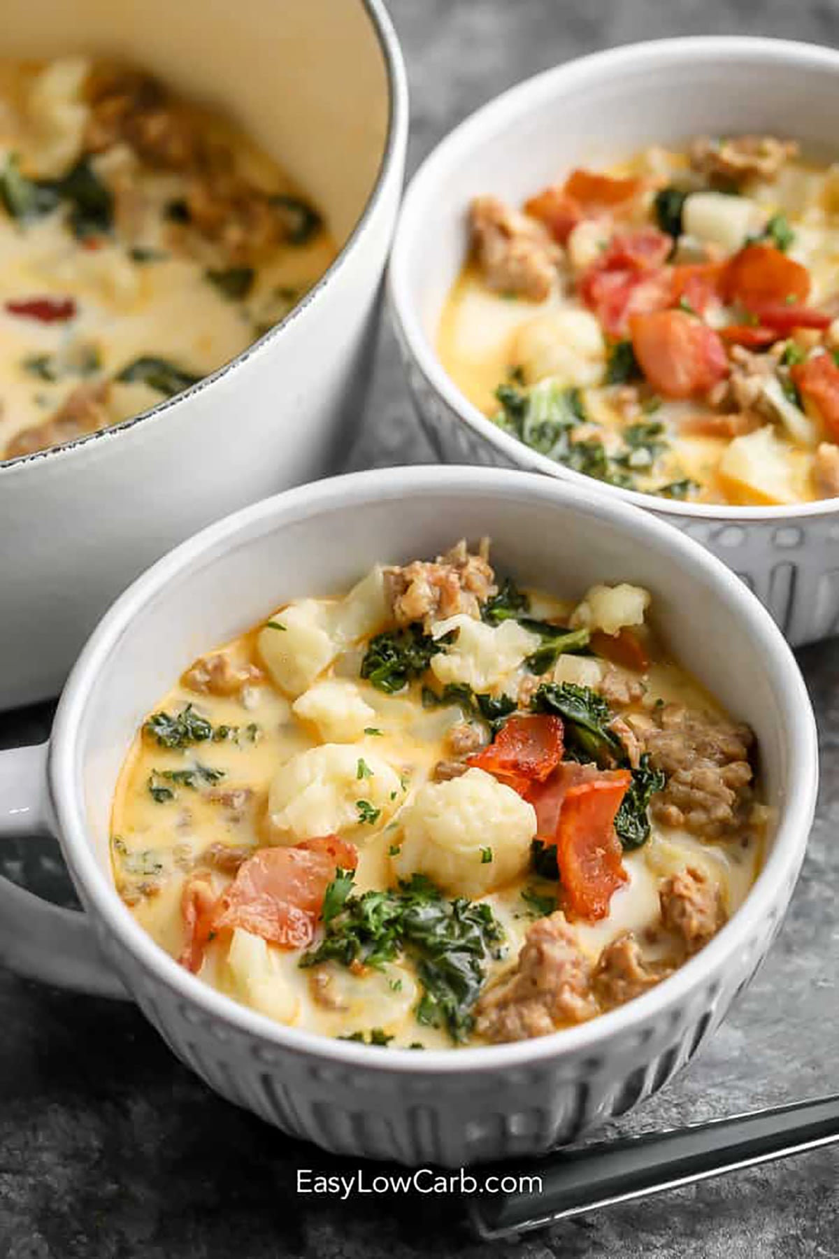 Keto Zuppa Toscana is a creamy, hearty soup filled with Italian sausage, bacon, cauliflower, and kale. The rich chicken broth and cream create a comforting base with plenty of savory flavor in every spoonful. This easy low carb soup makes a satisfying dinner for chilly days and is a delicious way to enjoy Italian-inspired comfort food without the potatoes. Top each bowl with crispy bacon and Parmesan. Try this cozy keto soup tonight. #easylowcarb bowls of Keto Zuppa Toscana with bacon.